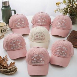 May include: A collection of seven baseball caps in shades of pink and cream, each featuring white embroidered text that reads GLAMP MIKEL and Smoke Show in the SMOKE MOUNTAINS, accompanied by a small graphic of mountain peaks and pine trees.