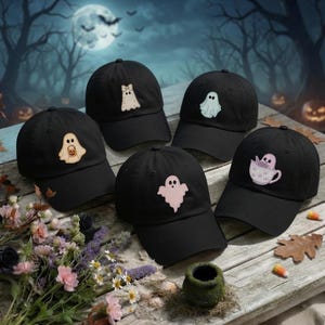 May include: Five black baseball caps arranged on a wooden surface, each featuring a unique embroidered ghost design. The designs include a ghost with a bow, a blue ghost, a ghost holding a pumpkin, a pink ghost, and a ghost inside a teacup.