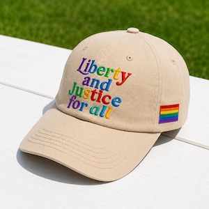 May include: A beige baseball cap featuring the embroidered text Liberty and Justice for all in multi-colored letters on the front panel. A small rectangular patch with a horizontal rainbow stripe is stitched onto the side of the cap.
