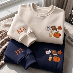 May include: A cream sweatshirt with the embroidered letters MA and a navy sweatshirt with the embroidered letters DT. Both feature embroidered designs of a coffee mug with a heart, a book, a candle, and an orange pumpkin on the chest area.