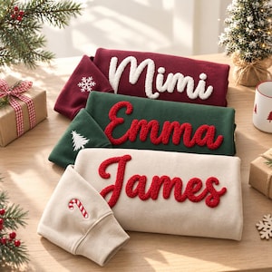May include: Three folded sweatshirts in maroon, dark green, and beige. The maroon sweatshirt features the white text Mimi and a snowflake. The green sweatshirt has the red text Emma and a tree. The beige sweatshirt has the red text James and a candy cane.