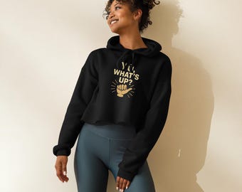 Op de afbeelding: Zwarte cropped hoodie met gouden tekst "Yo, What's Up?" en een handgebaar. De hoodie heeft een capuchon met trekkoord en lange mouwen. Het model draagt een blauwgrijze fietsbroek.