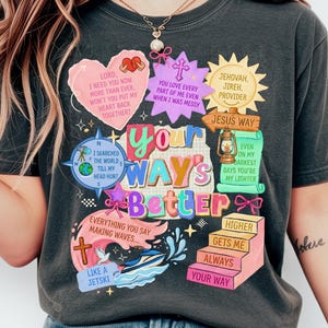 May include: A charcoal gray t-shirt featuring a colorful graphic print with the text "YOUR WAY'S BETTER" at the center. The design includes various religious-themed illustrations, including a heart, a cross, a compass, a lantern, and stairs.