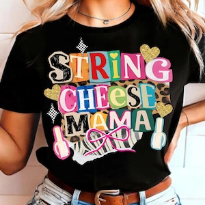 May include: Black t-shirt featuring the text "STRING CHEESE MAMA" in a colourful, layered design. The graphic includes gold glitter hearts, nail polish bottle illustrations, and a pink bow. The shirt has a crew neck and short sleeves.