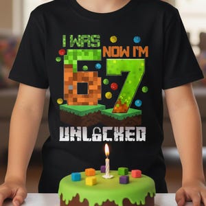 Puede incluir: Camiseta negra con el texto "I WAS 6, NOW I'M 7 UNLOCKED" en fuente pixelada. Una tarta de cumpleaños con glaseado verde y decoraciones cuadradas coloridas está frente a la camiseta. Una vela encendida está encima de la tarta.
