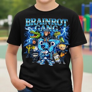 以下が含まれることがあります： 黒いTシャツ。青色の「BRAINROT GANG」の文字と稲妻のグラフィック。ワニ、猿、カエル、サメなどの漫画のキャラクターが描かれています。柔らかい素材で作られたTシャツです。