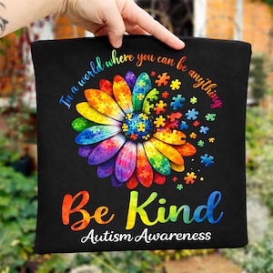 May include: Black square pillow with a colorful daisy design made of puzzle pieces. The text "In a world where you can be anything" and "Be Kind Autism Awareness" are also printed on the pillow.