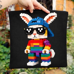 May include: Black tote bag featuring a colorful Lego-style bunny wearing sunglasses and a blue cap. The bunny is constructed with rainbow-colored blocks and has white ears with orange accents. The bag is held up by a hand.