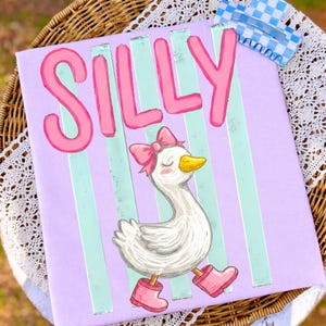 Könnte beinhalten: Ein Leinwandkunstwerk mit dem Wort "SILLY" in rosa Buchstaben. Darunter ist eine Cartoon-Gans mit rosa Schleife und Stiefeln abgebildet. Der Hintergrund ist Lavendel mit vertikalen mintgrünen Streifen. Oben ist eine blau-weiß karierte Klammer befestigt.