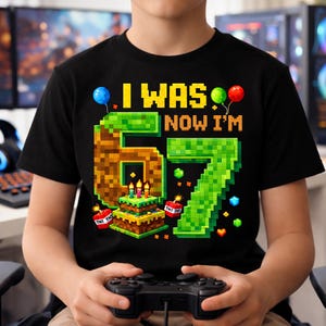Puede incluir: Camiseta negra con un diseño pixelado para un sexto cumpleaños. La camiseta presenta el texto "I WAS 6 NOW I'M 7" con un "6" y un "7" pixelados, una tarta de cumpleaños, globos y bloques de TNT. La camiseta la lleva una persona que sostiene un mando de videojuegos.