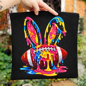 May include: Black tote bag featuring a colorful design of a football with bunny ears. The football is red with white stripes and laces, and the bunny ears and dripping paint are in shades of pink, yellow, blue, and orange.