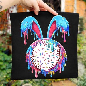 Puede incluir: Cojín cuadrado negro con un diseño de béisbol con orejas de conejo azules y glaseado goteando colorido. La pelota de béisbol está cubierta de chispas y las orejas son azules con el interior rosa.