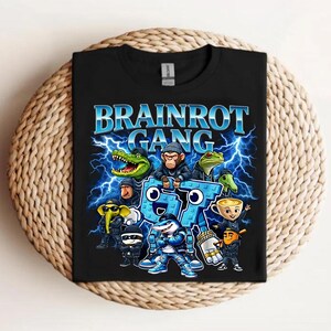 以下が含まれることがあります： 稲妻模様の青い文字で「BRAINROT GANG」と書かれた黒いTシャツ。グラフィックには、ワニ、猿、カエルなどの漫画のキャラクターが含まれています。Tシャツは織られた表面に折りたたまれています。