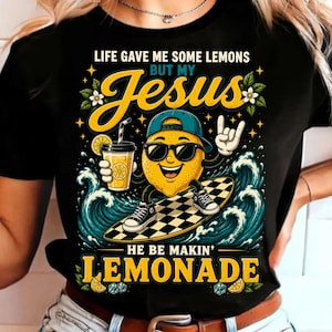 Può includere: Una t-shirt nera con la grafica di un limone cartoon che indossa occhiali da sole e un berretto blu, mentre fa surf su una tavola a scacchi tra onde blu. Il testo recita: LIFE GAVE ME SOME LEMONS BUT MY JESUS HE BE MAKIN' LEMONADE.