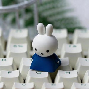 May include: A small, white, rabbit-shaped figurine with two upright ears, black dot eyes, and a black cross-shaped mouth, wearing a solid dark blue dress. The figurine is positioned on top of a white computer keyboard with green lettering.