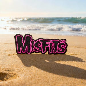 以下が含まれることがあります： 砂浜に置かれた、ピンク色の「Misfits」の文字が刺繍された黒いワッペン。背景には海が広がり、ワッペンはピンク色の縁取りがあり、砂の上に影を落としています。
