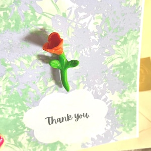 Peut inclure: Carte de remerciement faite à la main avec une fleur 3D orange et verte. La carte présente un motif floral vert et lavande et une étiquette blanche en forme de nuage avec les mots "Thank you" en noir.