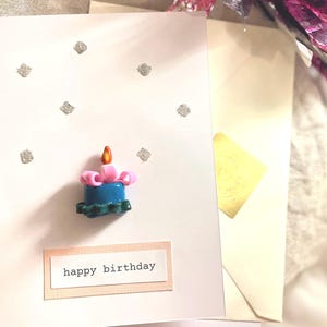 Peut inclure: Carte de vœux blanche avec un motif de bougie en 3D. La bougie est bleue avec un nœud rose et une flamme jaune. La carte porte l'inscription "happy birthday" et est ornée d'accents de paillettes argentées.