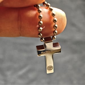 May include: A silver-colored cross pendant with a ball chain necklace. The cross has a polished finish and is engraved with the words "By Thy Will" and a circular design at the bottom. The necklace is held by a finger.