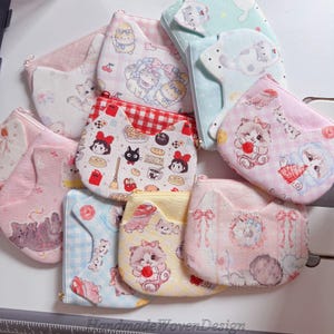 May include: A collection of small, fabric pouches in various pastel colors, featuring cat ear designs and whimsical illustrations. The pouches showcase patterns like gingham, floral, and character prints, with zipper closures and a handmade aesthetic.