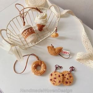 May include: A collection of handmade crochet food-themed keychains, including a coffee cup, ice cream cone, fortune cookie, donut, and chocolate chip cookies. Each keychain is crafted with detailed stitching and features a leather strap for attachment.