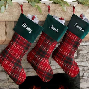 Könnte beinhalten: Drei Weihnachtsstrümpfe mit rot-grünem Karomuster. Jeder Strumpf hat eine grüne Samtmanschette mit einem weißen Namen: Hailey, Jacob und Cameron. Sie hängen an einem Holzkamin.