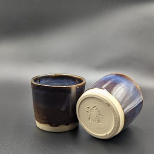 May include: Two ceramic cups with a dark blue and brown glaze. The cups have a white base and a cylindrical shape. One cup is upright, and the other is tilted, revealing a stamped symbol on the bottom.