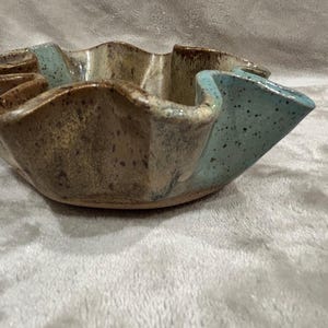 May include: A ceramic bowl with a wavy, scalloped rim featuring a speckled glaze. The exterior and interior surfaces display a blend of earthy brown tones with dark speckles, contrasted by sections of light blue glaze with dark speckles.