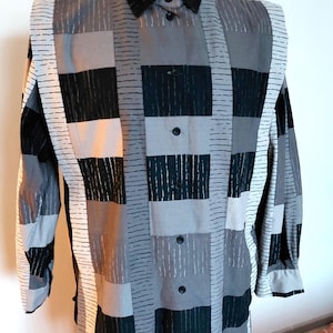 Puede incluir: Camisa de manga larga con un diseño de patchwork en tonos grises, negros y blancos. La camisa tiene un cuello negro y una botonadura. El patrón incluye cuadrados y rayas verticales.