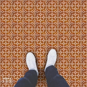 K&ouml;nnte beinhalten: Draufsicht auf einen gemusterten Boden mit einem sich wiederholenden floralen Design in Braun- und Wei&szlig;t&ouml;nen. Wei&szlig;e Turnschuhe und blaue Jeans sind am unteren Bildrand zu sehen, was darauf hindeutet, dass eine Person auf dem Boden steht.