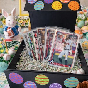 May include: A black box decorated with colorful Easter egg stickers, filled with baseball cards and shredded paper. A baseball-themed Easter display with a bunny, baseballs, and a miniature helmet.