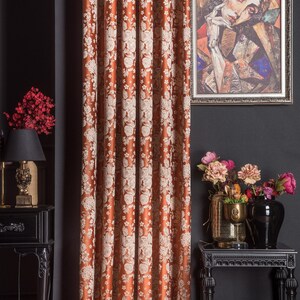Pu&ograve; includere: Tenda con motivo floreale color ruggine. La tenda presenta un motivo ripetuto di fiori bianchi. Un ritratto incorniciato &egrave; appeso alla parete scura. &Egrave; visibile anche un tavolino nero con una lampada e un vaso di fiori.