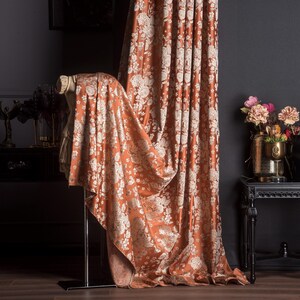 Pu&ograve; includere: Tenda con motivo floreale color ruggine e tessuto drappeggiato abbinato. La tenda &egrave; accanto a un tavolo nero con un vaso di fiori. La stanza ha pareti scure e pavimento in legno. Elementi decorativi: vasi e un tavolino.