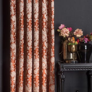 Pu&ograve; includere: Tenda color ruggine con motivo floreale color crema. Il tessuto &egrave; drappeggiato in pieghe verticali, creando un aspetto strutturato. Un tavolino nero con vasi di fiori &egrave; accanto.
