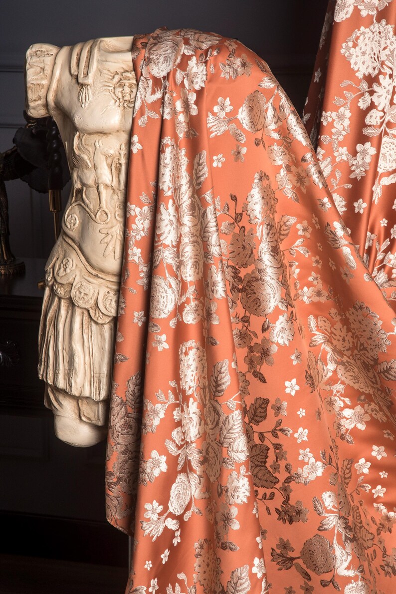 Pu&ograve; includere: Un tessuto drappeggiato color ruggine con un motivo floreale nei toni del bianco e dell'argento. Il tessuto &egrave; drappeggiato su un busto classico. Il design floreale comprende rose e altri fiori, creando un tessuto lussuoso.
