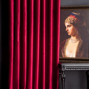 Può includere: Una tenda di velluto rosso intenso, con pieghe verticali, occupa gran parte dell'immagine. Un ritratto incorniciato di una donna, posto su un tavolo nero, è parzialmente visibile, creando un contrasto sorprendente.