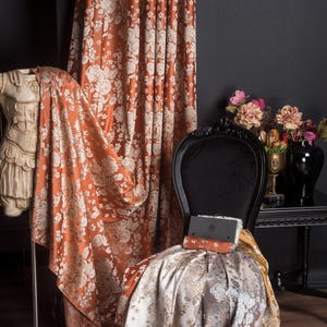Pu&ograve; includere: Tessuto per tende con motivo floreale color ruggine drappeggiato su una sedia nera. Il tessuto &egrave; drappeggiato anche su una statua e un rotolo di tessuto &egrave; sulla sedia. Un tavolo nero con fiori &egrave; sullo sfondo.