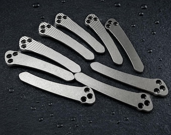 May include: A collection of seven curved metal pocket clips, each featuring a three-hole mounting pattern at one end. Four clips display a fine linear grooved texture on their surface, while three clips have a smooth, matte metallic finish.