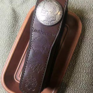 Puede incluir: Una funda Leatherman de cuero marr&oacute;n con un adorno de moneda de plata. La funda presenta dise&ntilde;os en relieve de inspiraci&oacute;n nativa americana y la marca Leatherman. La moneda es circular con un dise&ntilde;o de calavera.