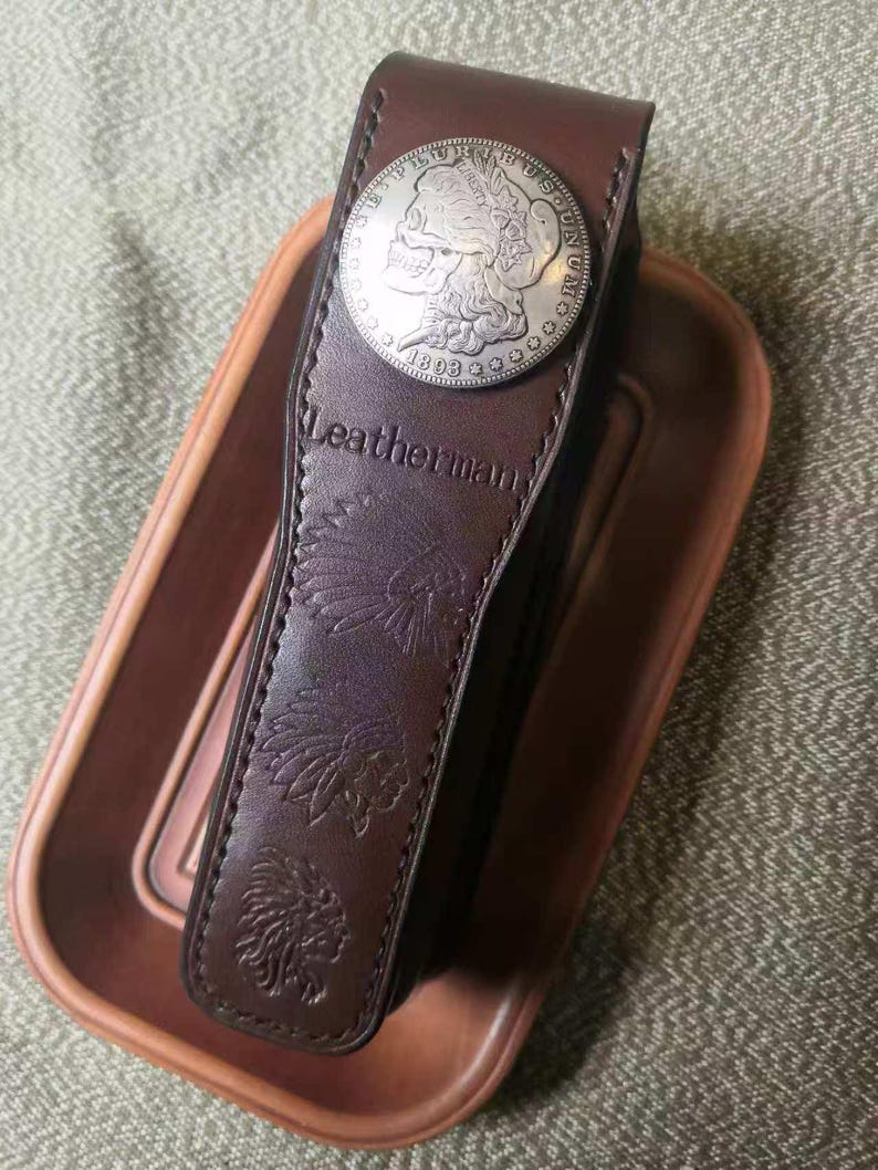 Puede incluir: Una funda Leatherman de cuero marr&oacute;n con un adorno de moneda de plata. La funda presenta dise&ntilde;os en relieve de inspiraci&oacute;n nativa americana y la marca Leatherman. La moneda es circular con un dise&ntilde;o de calavera.
