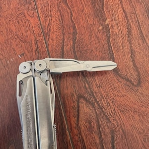 Peut inclure: Un outil multifonction Leatherman Wave argenté avec des ciseaux déployés, posé sur une surface en bois. L'outil est partiellement ouvert, révélant ses différents composants. Le texte "LEATHERMAN WAVE" est visible.