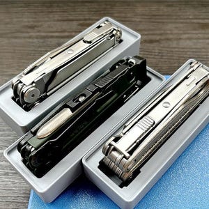 Könnte beinhalten: Drei Multitools in grauen Boxen. Die Werkzeuge sind silber und schwarz, mit verschiedenen Klappkomponenten. Die Boxen sind grau und rechteckig, und der Hintergrund ist eine Mischung aus Holz und Blau.