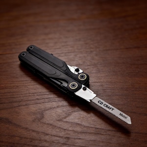 May include: A black multi-tool with a partially extended saw blade. The blade is silver with the text "CD CRAFT Metal" printed on it. The tool is lying on a dark brown wooden surface.