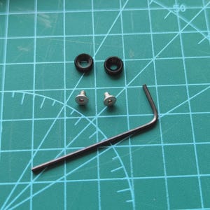 May include: A selection of small hardware components. The image shows two black rubber washers, two silver screws, and a dark metal Allen key. The items are arranged on a green grid.