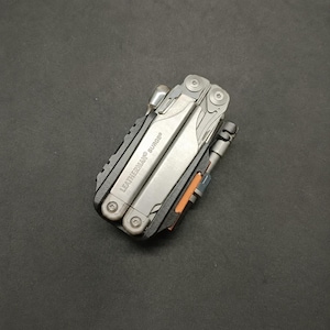 May include: A silver and black Leatherman Surge multi-tool in a folded, closed position. The metal body features the text LEATHERMAN SURGE engraved on the side. The tool includes visible screws, a black frame, and a small orange accent detail.