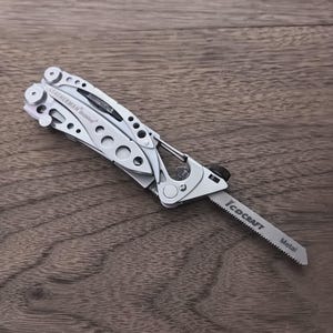 May include: A silver multi-tool with a saw blade extended. The tool has the brand name "LEATHERMAN" and the text "Tread" printed on it. The saw blade has the text "TCD CRAFT" and "Metal" printed on it. The tool is on a wooden surface.