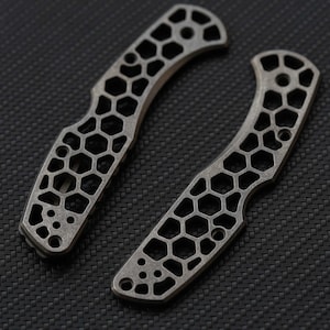 May include: Two grey metal knife handle scales featuring a cut-out honeycomb pattern. Each piece has a curved shape with multiple circular screw holes positioned at the ends and along the body, showcasing a matte, textured finish throughout.