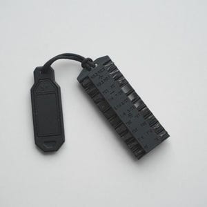 Puede incluir: Calibrador de paso de rosca de metal negro con forma rectangular y dientes dentados. El calibrador tiene texto que indica los tamaños de rosca y una pequeña etiqueta con un cordón adjunto. Fondo blanco.