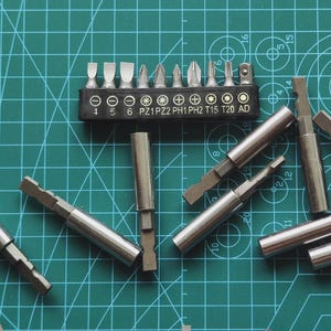 以下が含まれることがあります： 緑色のカッティングマットの上に、銀色のドライバービットと黒いホルダーが配置されています。ビットは、マイナス、プラス、星型など、さまざまなヘッド形状をしています。ホルダーには数字と文字が記載されています。