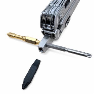 May include: A silver multi-tool with a textured handle, featuring a central metal block holding a gold-colored screwdriver bit and a silver-colored screwdriver bit. A separate black screwdriver bit is positioned below the multi-tool.